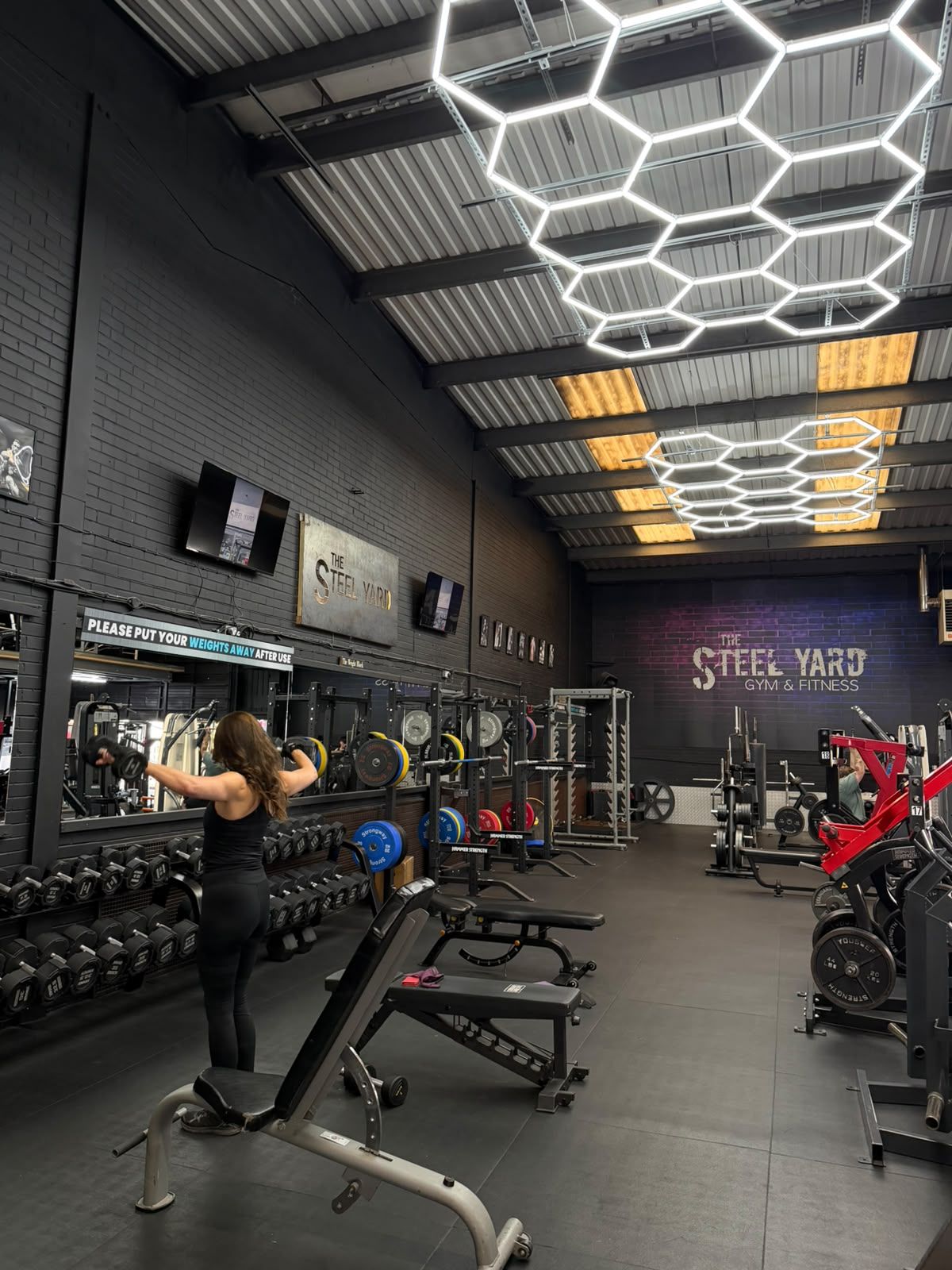 A member training in the free-weights area, beneath hexagonal lights and The Steel Yard wall sign.